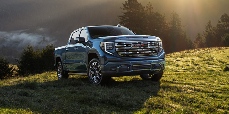 Silver GMC Sierra 1500 positioned on a grassy terrain with a mountainous landscape and misty sunrise backdrop.